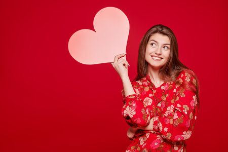 Smiling young woman in red dress holds a red heart and smiles. Studio portrait on a red background...