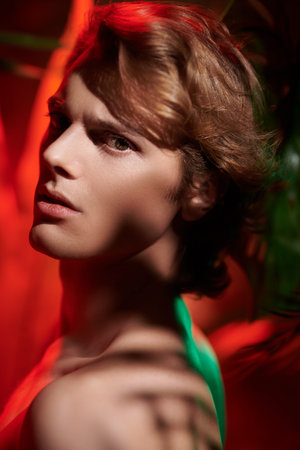 Handsome young man fashion model with naked torso posing among tropical plants in red light. Men's...