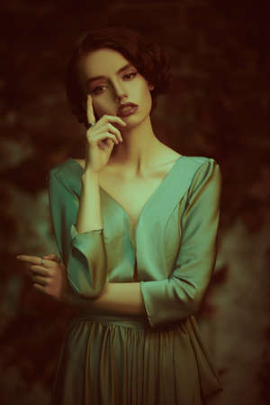 Beautiful pensive girl in an elegant dress stands against the background of an old vintage wall. Art fashion. Hair and makeup in the style of the 20s.の写真素材
