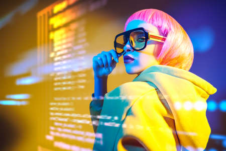 Beauty trends. Attractive fashion model girl with bright makeup and pink hair poses in modern sunglasses and yellow suit against the background of holographic data. Information technology.の写真素材