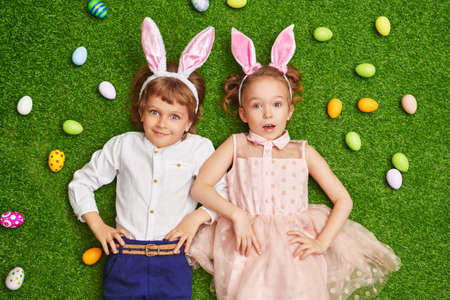 Cute smiling boy and girl with bunny ears are lying on a green lawn with colorful eggs. Easter holiday.の写真素材