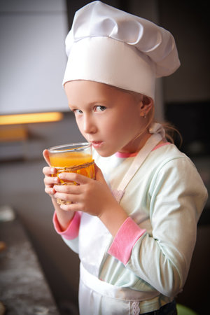 Portrait of a cute little girl in apron and chef's hat secretly drinks orange juice. Fresh juice and healthy eating.の写真素材