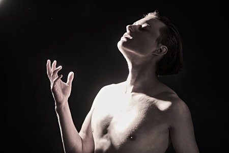 Handsome emotionalman poses shirtless in the dark, turning and aiming for the light. Black...