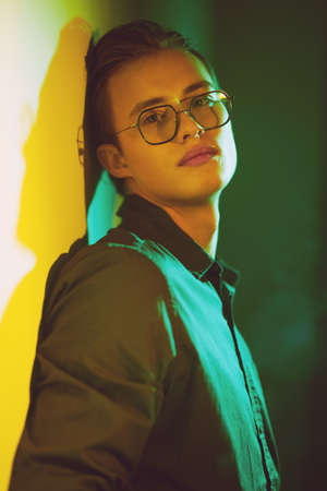 Handsome young man stands, sad and alone, leaning against the wall in dark green and yellow lighting.の写真素材