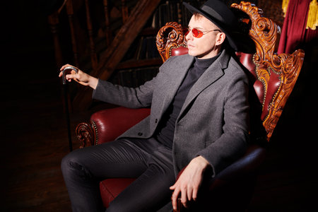 Imperious mature man in stylish expensive clothes and glasses sits in the armchair in the luxury...