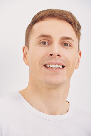 Positive handsome man stands leaning against a white wall and smiles. Athletic man posing at studio....