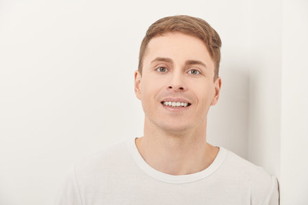 Positive handsome man stands leaning against a white wall and smiles. Men's health.