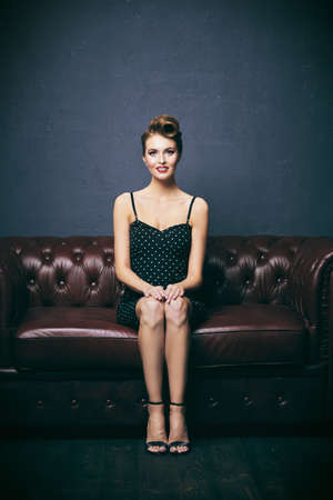 Pretty woman with pin-up hairstyle and make-up is posing on a leather sofa. Pin-up beauty and fashion concept.の写真素材