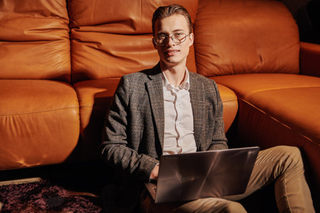 Well-groomed young man in elegant clothes and glasses sits by a leather sofa with laptop in a modern...
