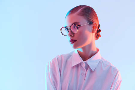 Portrait of a beautiful young woman in elegant glasses and white shirt on light blue background in pink lighting. Copy space. Business style. Glasses fashion.の写真素材