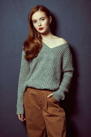 Young attractive girl in fashionable modern clothes stands leaning against the gray wall. Fashion...