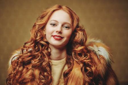 Beautiful young woman with long red hair poses in a luxurious fox fur coat on a golden vintage background. Winter beauty fashion. Fur coat style.の写真素材