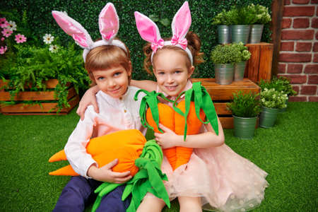 Joyful little kids with bunny ears sits with big carrots on a green lawn in a spring garden with Easter decorations. Easter holiday. Happy family.の写真素材