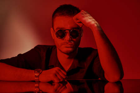 Modern stylish man in dark shirt and sunglasses posing with window shadows in red lighting. Men's fashion.の写真素材