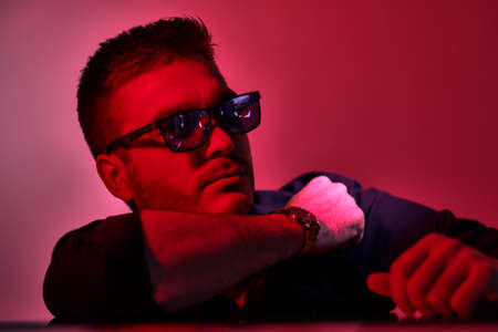 Modern stylish man in dark shirt and sunglasses posing in red and crimson lighting. Men's fashion.の写真素材