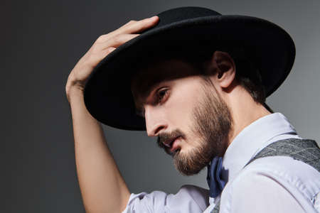 Close uo portrait of a handsome brunet man in elegant classic suit and black hat on a grey background. Men's beauty. Fashion.の写真素材