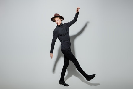 Handsome young man in black clothes and hat posing in motion at studio on a light gray background....