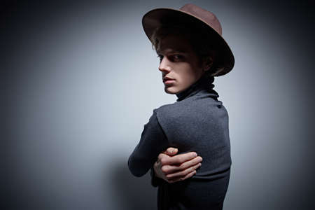 Attractive young man in a wide-brimmed hat and a turtleneck posing with his arm around his shoulders Fashion art shot on gray background with copy space.の写真素材