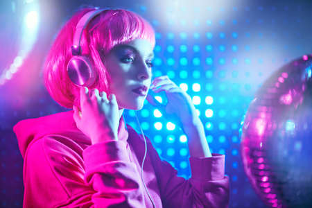 Disco girl. Attractive girl with bright glitter make-up and pink hair poses with disco ball and headphones in neon light. Night party. DJ girl.の写真素材