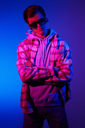 Modern young man in casual clothesand sunglasses posing at studio in mixed color light on blue...