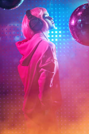 Disco girl with bright glitter make-up and pink hair poses with disco ball and headphones in smoky nightclub in neon light. Night party. DJ girl.の写真素材