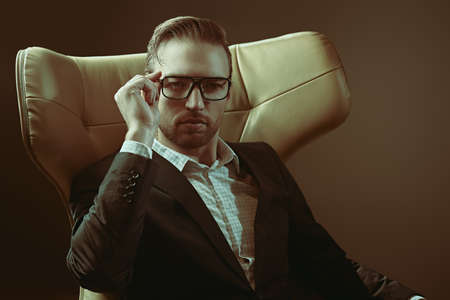 Portrait of a confident middle-aged businessman in elegant suit and glasses sitting in a leather chair and seriously looking at the camera. Success and leadership.の写真素材
