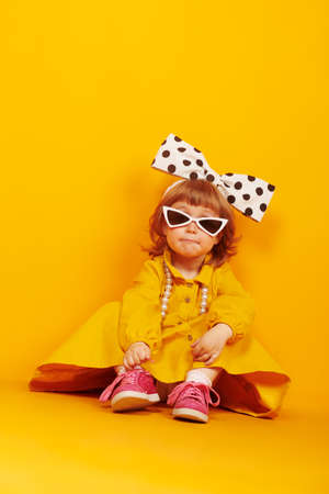 Lovely little child girl in fashionable dress, big bow and sunglasses poses on a bright yellow background. Kid's fashion.の写真素材