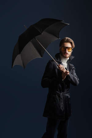 Men's fashion. Portrait of a handsome fashion model man posing at studio in a raincoat and with an umbrella on a dark blue background.の写真素材