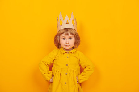 Cute little girl in a cardboard crown looks at the camera and smiles. Happy childhood. Kid's emotions and imagination. Studio portrait on a yellow background.の写真素材