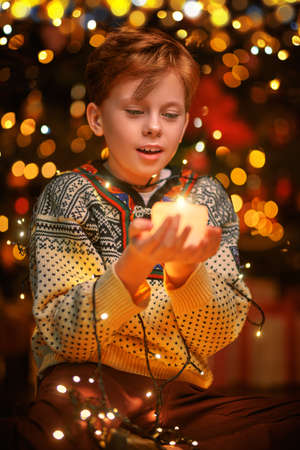 Portrait of a cheerful boy holding a burning candle candle and a glowing garland. Lights and sparkle is around. Merry Christmas and Happy New Year!の写真素材