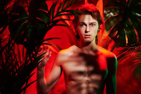 Athletic young man fashion model with naked torso poses among tropical plants. Men's beauty. Perfumery and beauty products. Jungle photoshoot.の写真素材