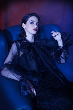 Fashion chic style. Beautiful brunette girl in fashionable black dress sits in a leather armchair in...