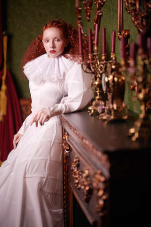 Portrait of as sophisticated lady with lush red hair with fine curls standing in a vintage interior in art dress with a ruffled renaissance collar. History of fashion and hairstyles.の写真素材