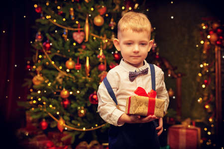 Dreams come true on Christmas. Happy little boy stands in front of a beautiful Christmas tree with a gift box and is smiling.の写真素材