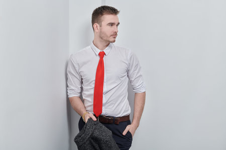 Handsome stylish man in white shirt and red tie poses near the white wall. Businessman portrait.