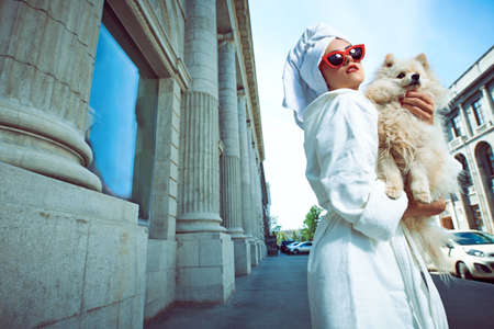 Glamorous lady in a white bathrobe with a white towel on her head and elegant red sunglasses walks along the street of a big city with a small dog in her arms. Beauty, fashion.の写真素材