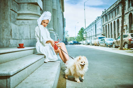 Stunning woman in a white terry dressing gown with a white towel on her head sitting on the stairs of building on a city street keeping the dog on a leash. Glamorous lifestyle. Fashion shot.の写真素材