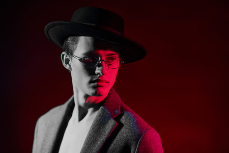 Handsome young man fashion model posing in a coat, a hat and glasses in crimson light gray lighting....