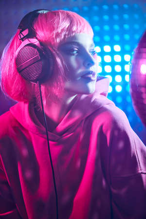 Disco girl. Attractive girl with bright glitter make-up and pink hair poses with disco ball and headphones in neon light. Night party. DJ girl.の写真素材