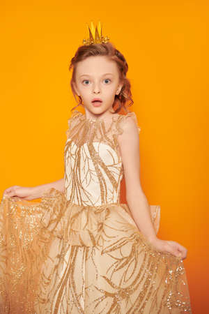 Emotional little princess girl in a golden dress and a crown looks at a camera and is surprised. Dreams and kid's fashion. Yellow background.の写真素材