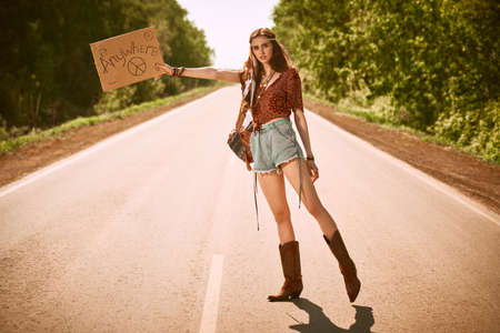 Beautiful hippie girl hitchhiking along a road. The spirit of freedom and adventures. Full length portrait.の写真素材