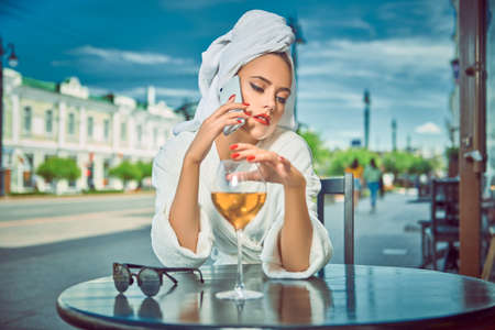 Attractive girl in a white terry dressing gown with a white towel on her head sits at a table in a street cafe with a glass of champagne and gossips on the phone. Fashion shot.の写真素材