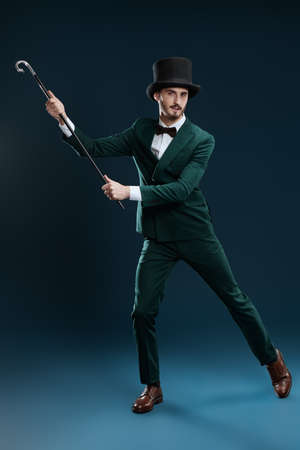 Elegant gentleman in suit and top hat dances with a cane. Studio portrait on a dark blue background. Men's fashion.の写真素材
