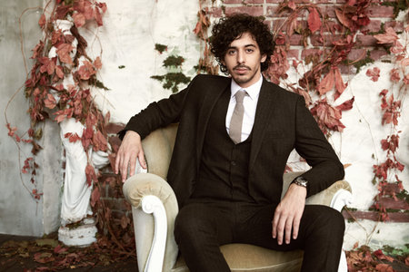 Handsome brunet man in elegant classic suit and a tie sits in an armchair on a vintage background....