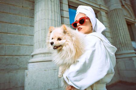 Gorgeous lady in a white bathrobe and elegant red sunglasses walks along the street of a big city with a small dog in her arms. Beauty, fashion.の写真素材