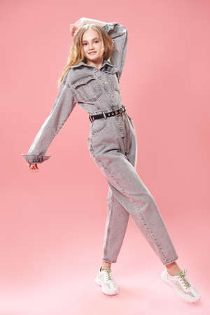 Full length portrait of a smiling girl teenager posing in fashionable denim overalls on a pink background. Studio fashion shot. Youth style.の写真素材