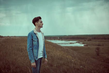 Male beauty, denim style. Portrait of a handsome young man in jeans jacket on the background of the green valley on a cloudy day.の写真素材