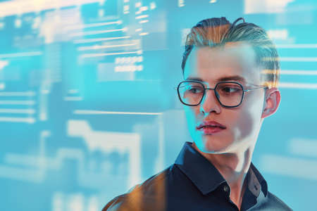 Business concept. Portrait of a handsome young man in elegant glasses on a blue background with digital holograms. Stock exchange.の写真素材