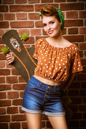 Pin-up girl posing by the brick wall with her skateboard. Pretty woman dressed in pin-up style, with...