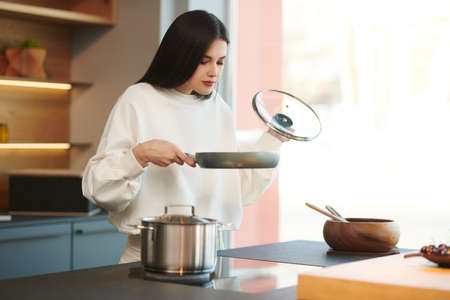 Pretty brunette girl stands with a pan in the kitchen and smiles. Healthy eating and diet. Modern interior, furniture for the kitchen.の写真素材